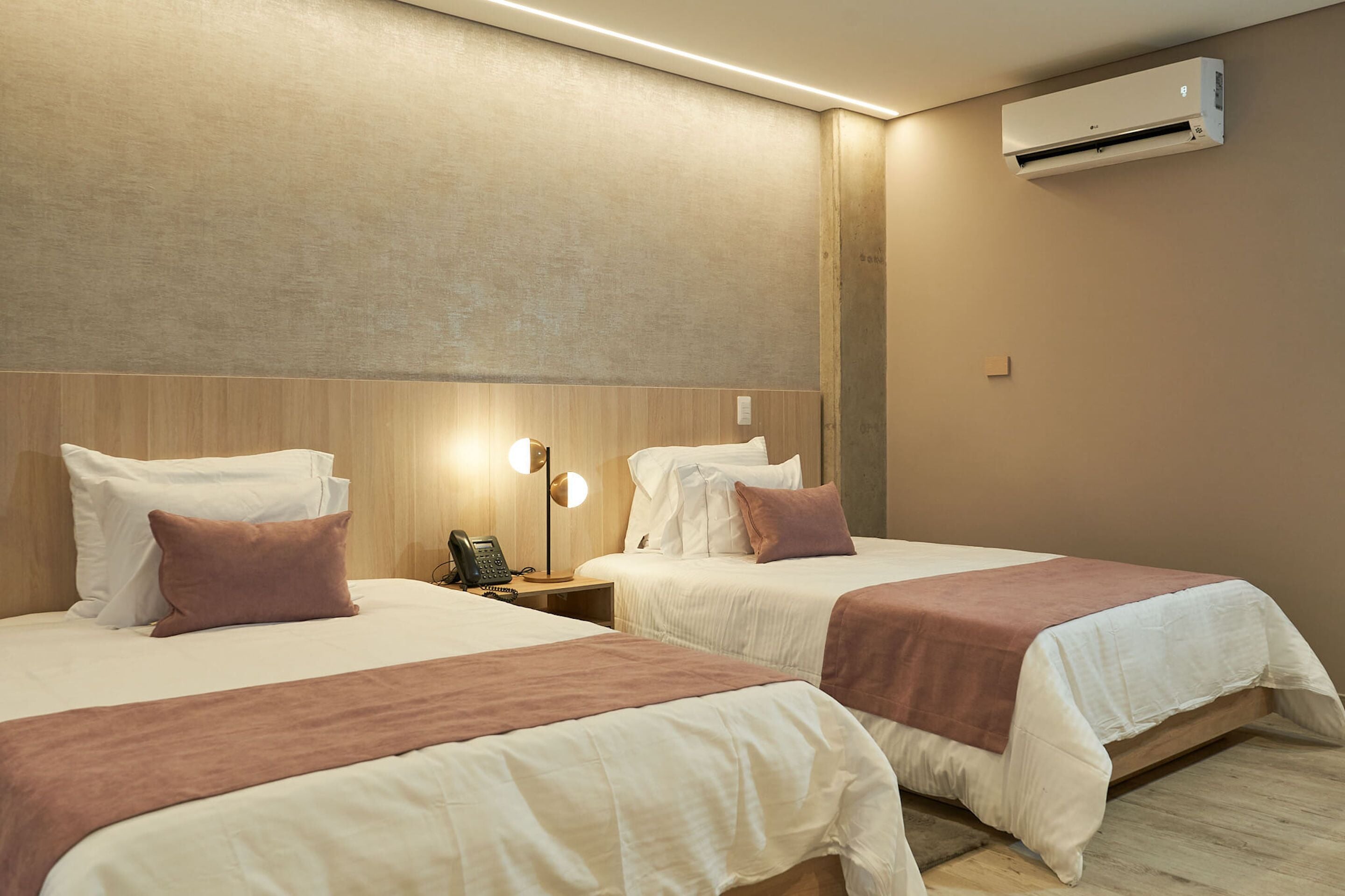 A hotel room with two beds, each with white bedding and a brown throw blanket. There is a wall-mounted air conditioner and a bedside table with a lamp and a phone.