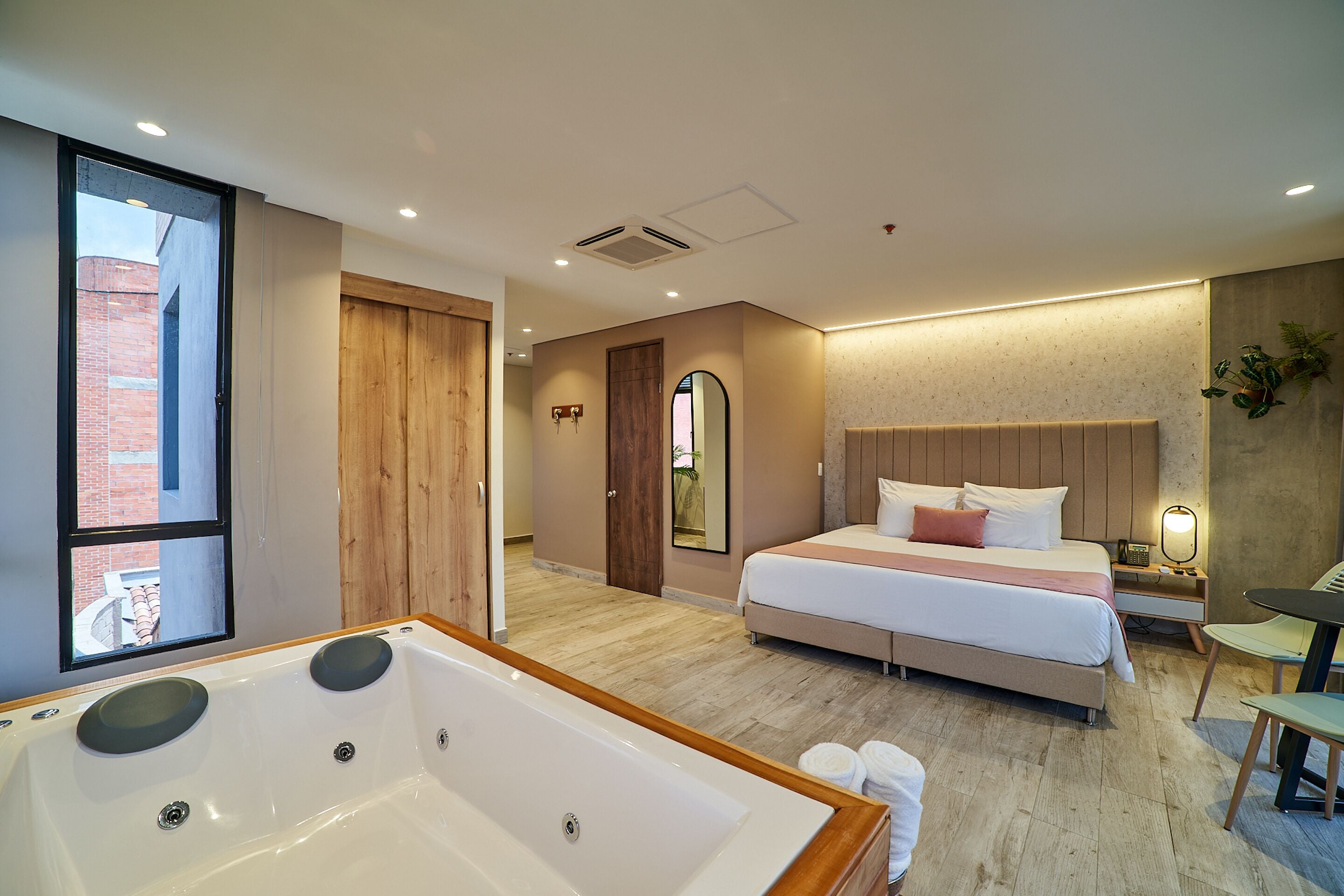 A modern hotel room with a large bathtub, a neatly made bed, and a view of a brick building through a window.