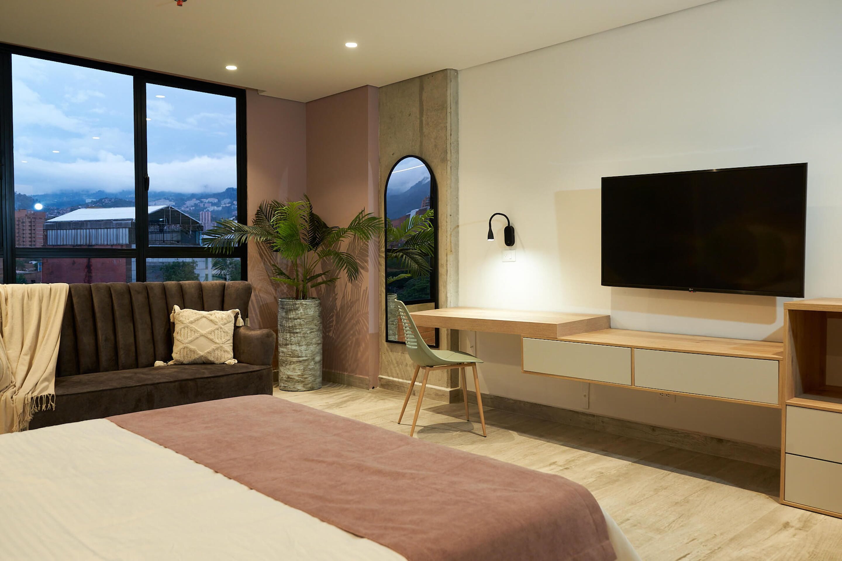 Un dormitorio moderno con una cama grande, un escritorio con televisor, vista a la ciudad y una planta en maceta.