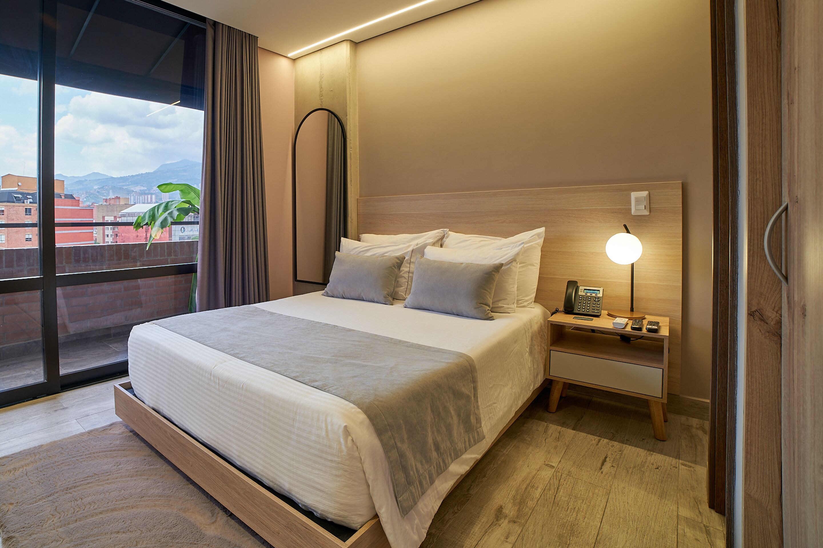A modern hotel room with a large bed, a bedside table with a lamp, and a view of buildings through a large window.
