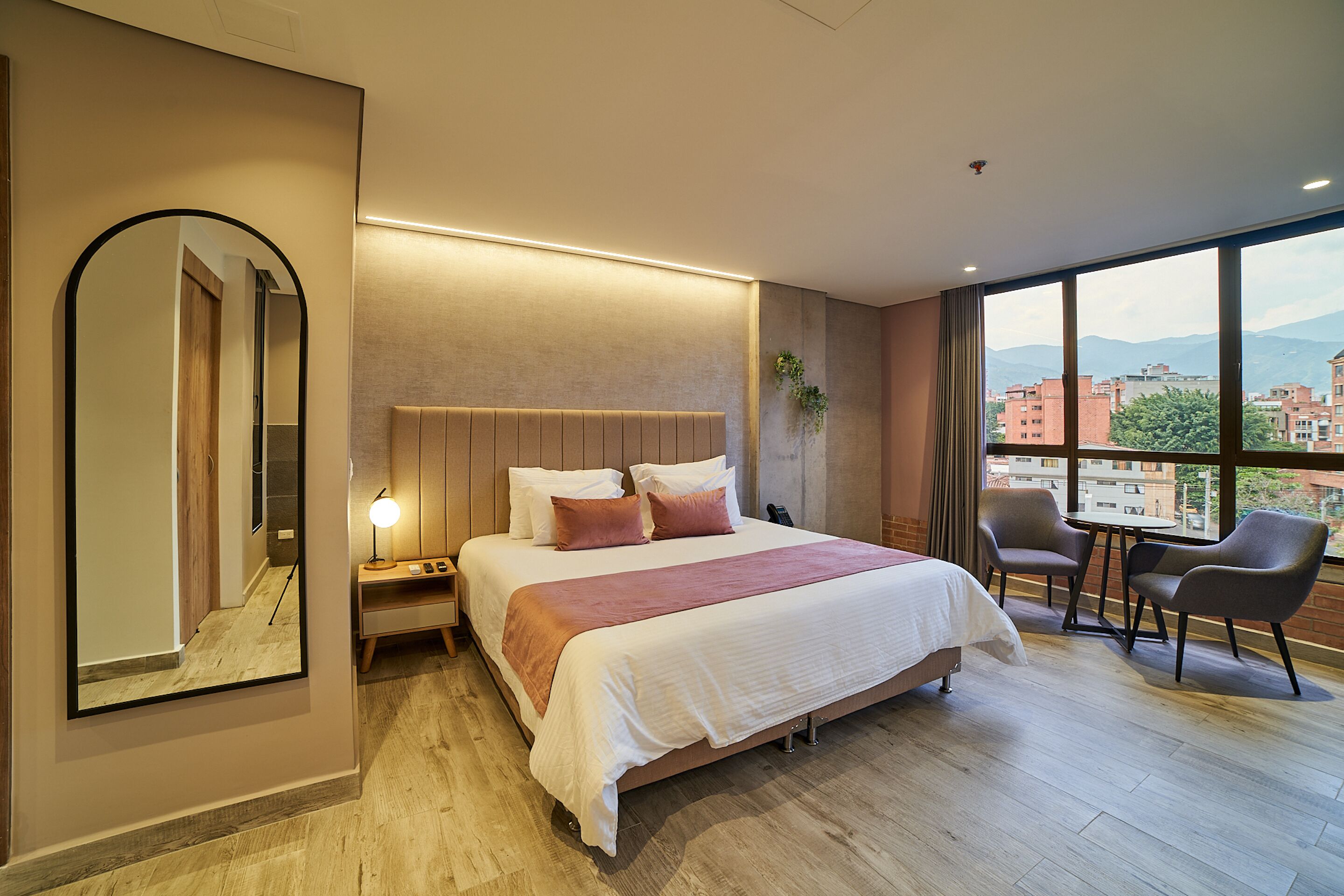 Habitación de hotel con una cama grande, un escritorio, una silla y vistas a edificios y montañas a través del ventanal.