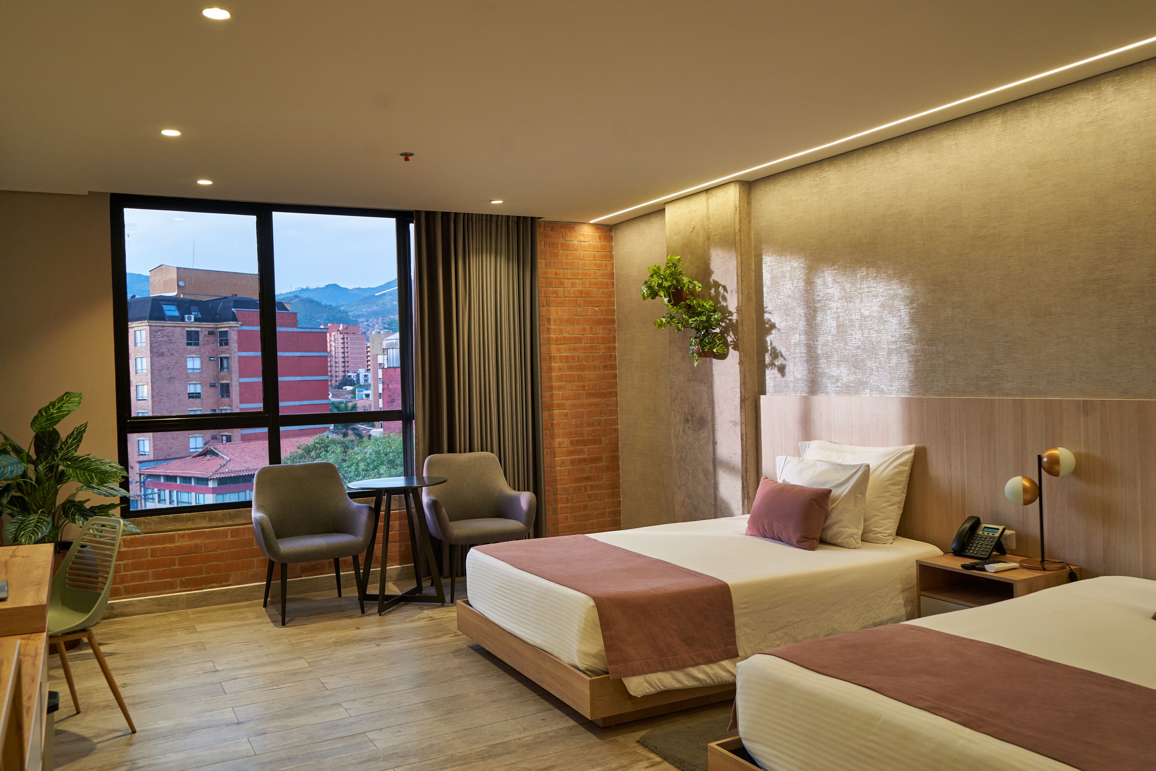 Ein Hotelzimmer mit zwei Betten, einem Schreibtisch, Stühlen, einer Zimmerpflanze und Blick auf Gebäude und Berge durch große Fenster.