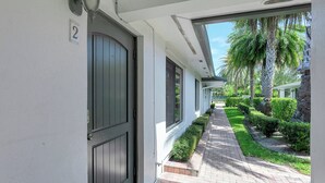 A corridor with a door marked '2', a pathway lined with hedges, and a view of palm trees and a building in the background. - Beautiful 1bd, prime location, free parking, bbq, pets (Delray Beach)