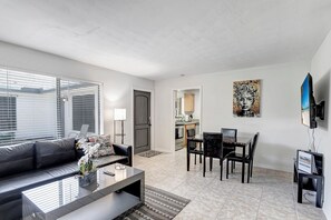 Uma sala de estar moderna com sofá, mesa de centro e área de jantar. Uma cozinha visível ao fundo, com uma televisão na parede.