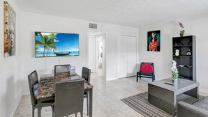 A modern living room with a dining area, white walls, a large painting of a beach, and a bookshelf with decorative items. - Beautiful 1bd, prime location, free parking, bbq, pets (Delray Beach)
