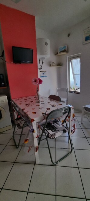 Un comedor con una mesa cubierta con mantel floreado, dos sillas de metal, un jarrón con flores, un televisor en la pared y una ventana con vistas.