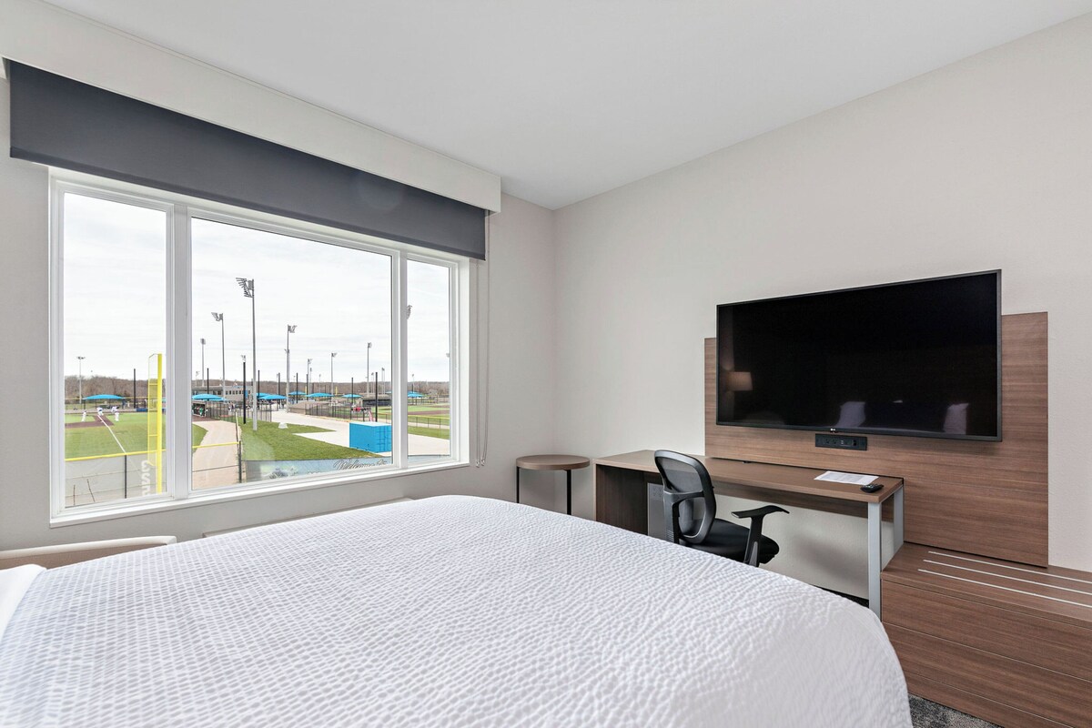 Habitación de hotel con una cama grande, un escritorio con silla, un televisor de pantalla plana montado en la pared y una ventana que ofrece vistas a un campo deportivo.