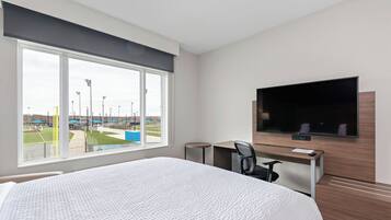 A hotel room with a large bed, a desk with a chair, a flat-screen TV mounted on the wall, and a window with a view of a baseball field.