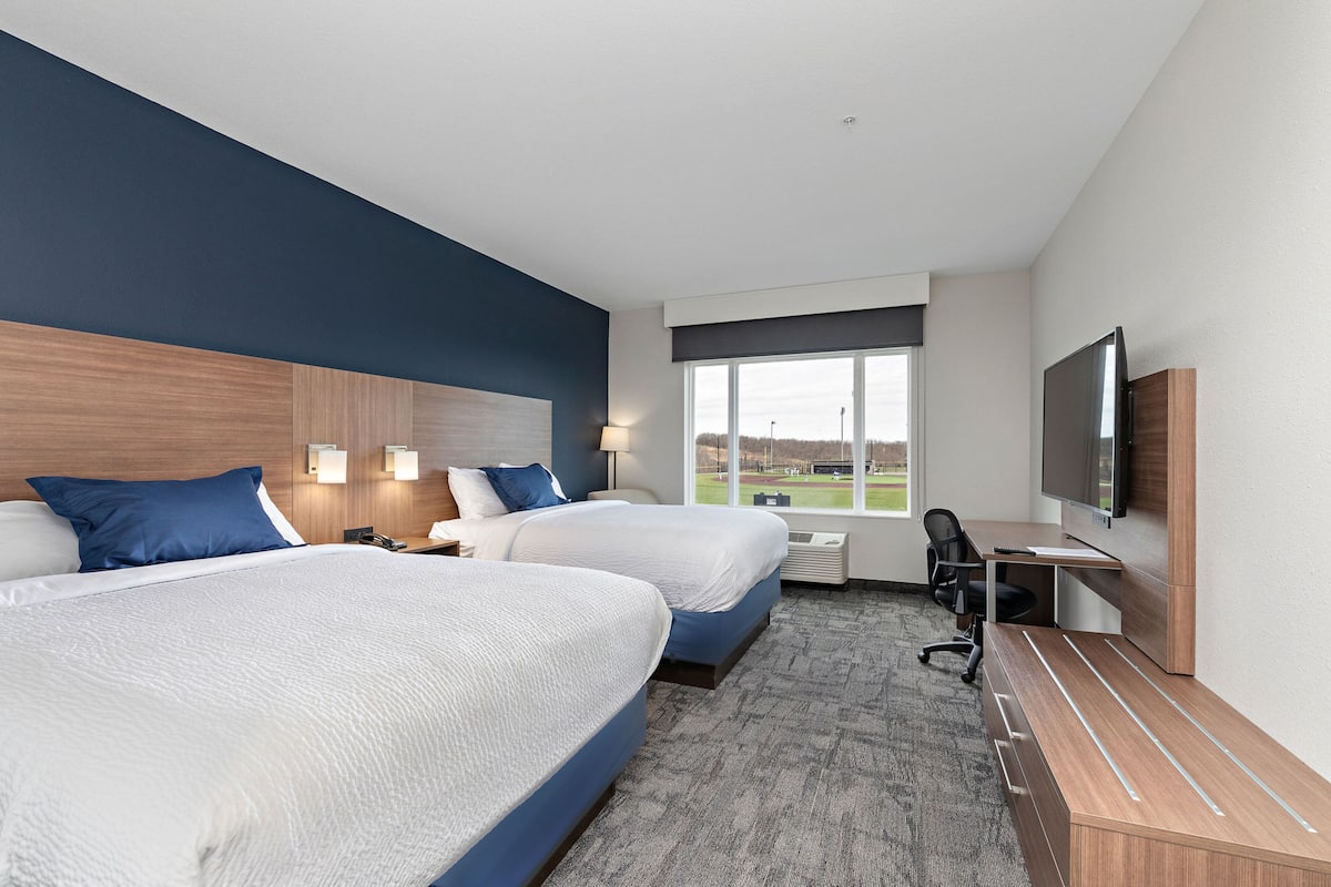 A hotel room with two beds, a desk, a TV, and a large window with a view of the outdoors.