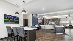 A modern, well-lit indoor space with a dining area, kitchen, and a large TV screen displaying a sports game. - Holiday Inn Express Kansas City North – Parkville by IHG (Parkville)