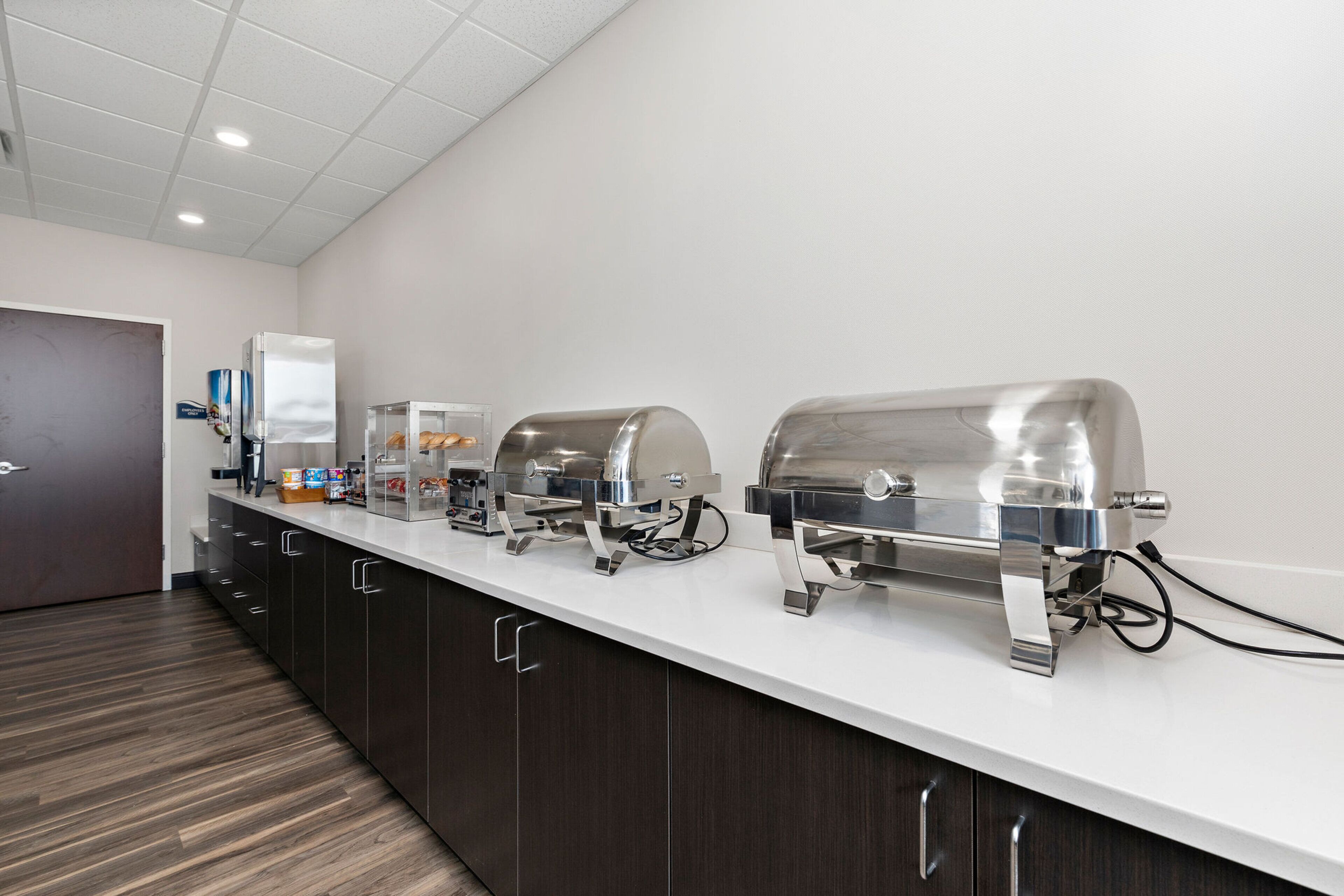 A buffet setup with three stainless steel chafing dishes on a white countertop, a refrigerator, and a coffee station.