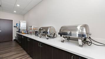 A buffet setup with three stainless steel chafing dishes on a white countertop, a refrigerator, and a coffee station.