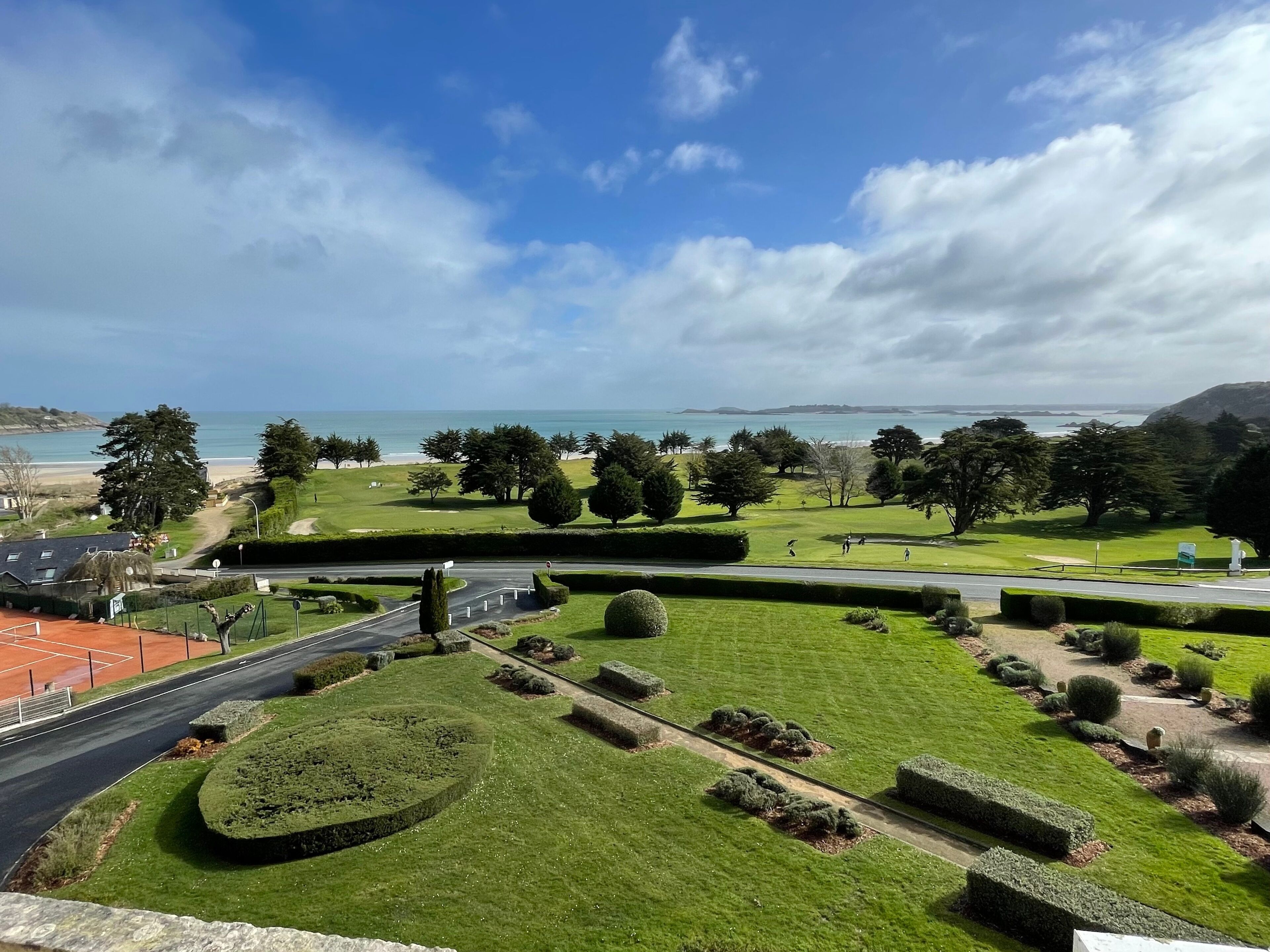 A golf course with well-manicured lawns, a road, and a view of the sea and hills in the distance.