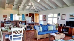 A spacious living room with a blue sofa, a dining table with chairs, and a television. - San Costanzo 2 br/ba Delightful breezes, Caribbean views, hot tub and generator! (St.John)
