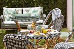 Un coin petit-déjeuner aménagé avec des chaises en osier, une table en verre et un canapé agrémenté de coussins à motifs, le tout dans un cadre de jardin.