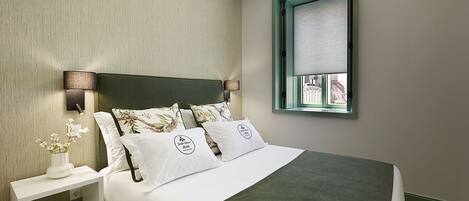 Quarto bem arrumado com cama, almofadas, mesa de cabeceira e janela com vista.