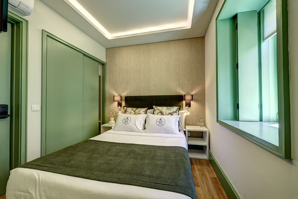 A modern hotel room with a large bed, a nightstand, a wall-mounted lamp, and a window with a view.