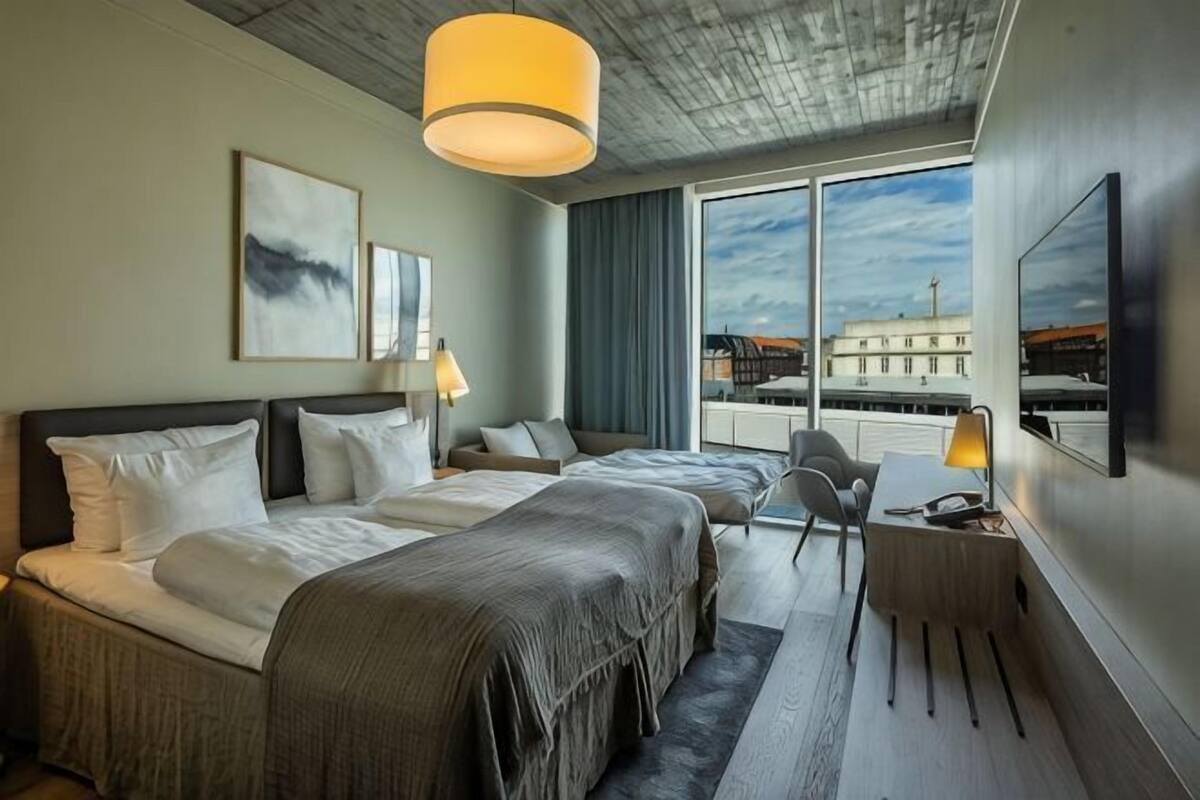 Une chambre d’hôtel avec deux lits, un bureau, une chaise, une télévision et une grande fenêtre donnant sur un immeuble.
