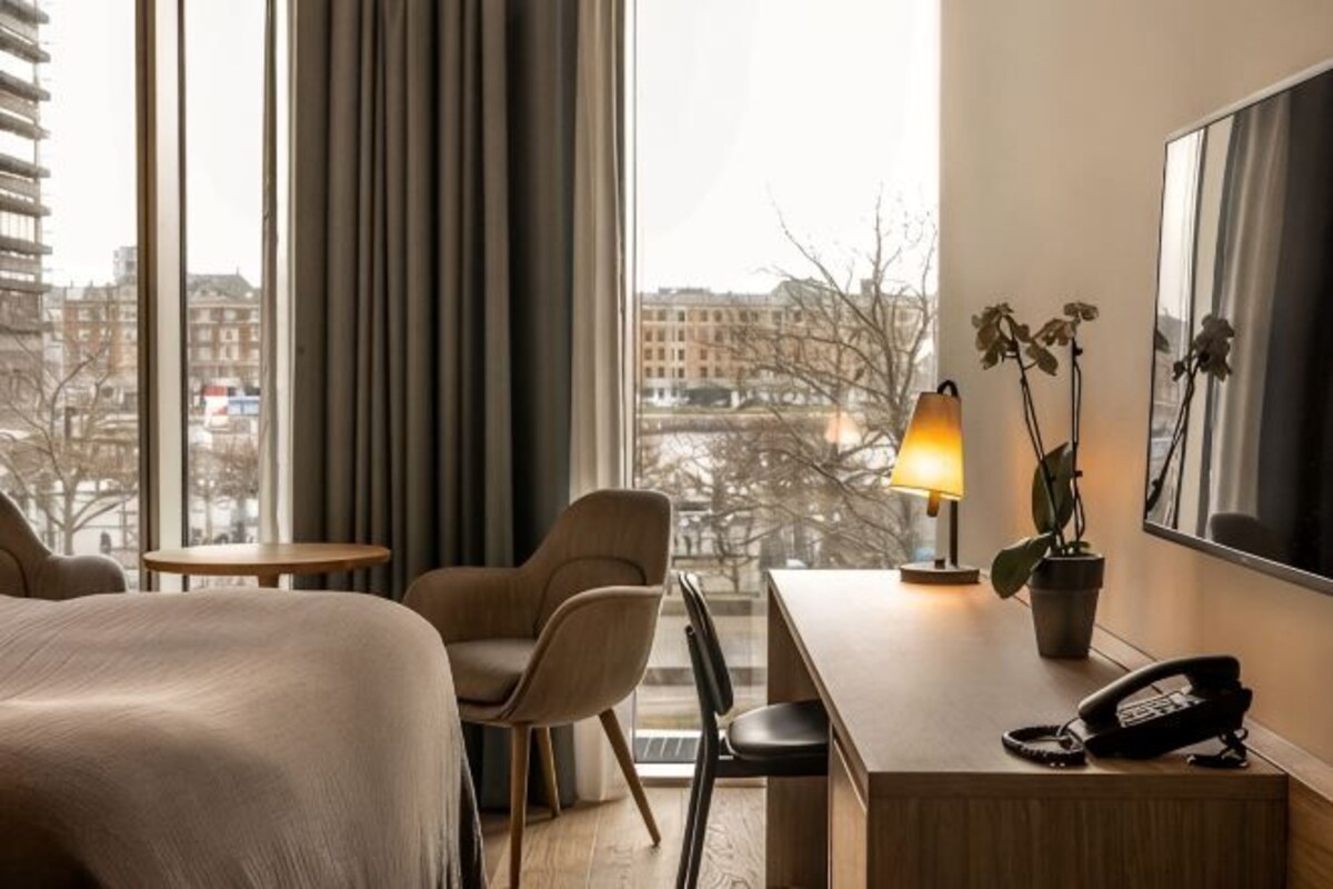 A hotel room with a bed, a desk, a chair, a lamp, a phone, and a view of buildings through large windows.