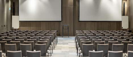 A conference room with rows of chairs facing two large projection screens, a podium, and a wooden panel wall.