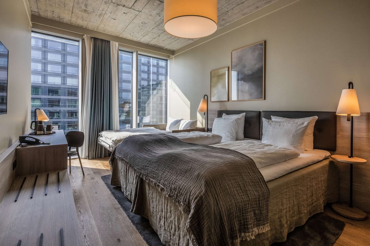 Une chambre d’hôtel moderne dotée d’un grand lit, de lampes de chevet, d’un bureau avec une chaise et offrant une vue sur la ville grâce à de grandes fenêtres.