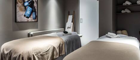 A spa room with two beds, a framed advertisement, a towel rack, and a shelf with towels.