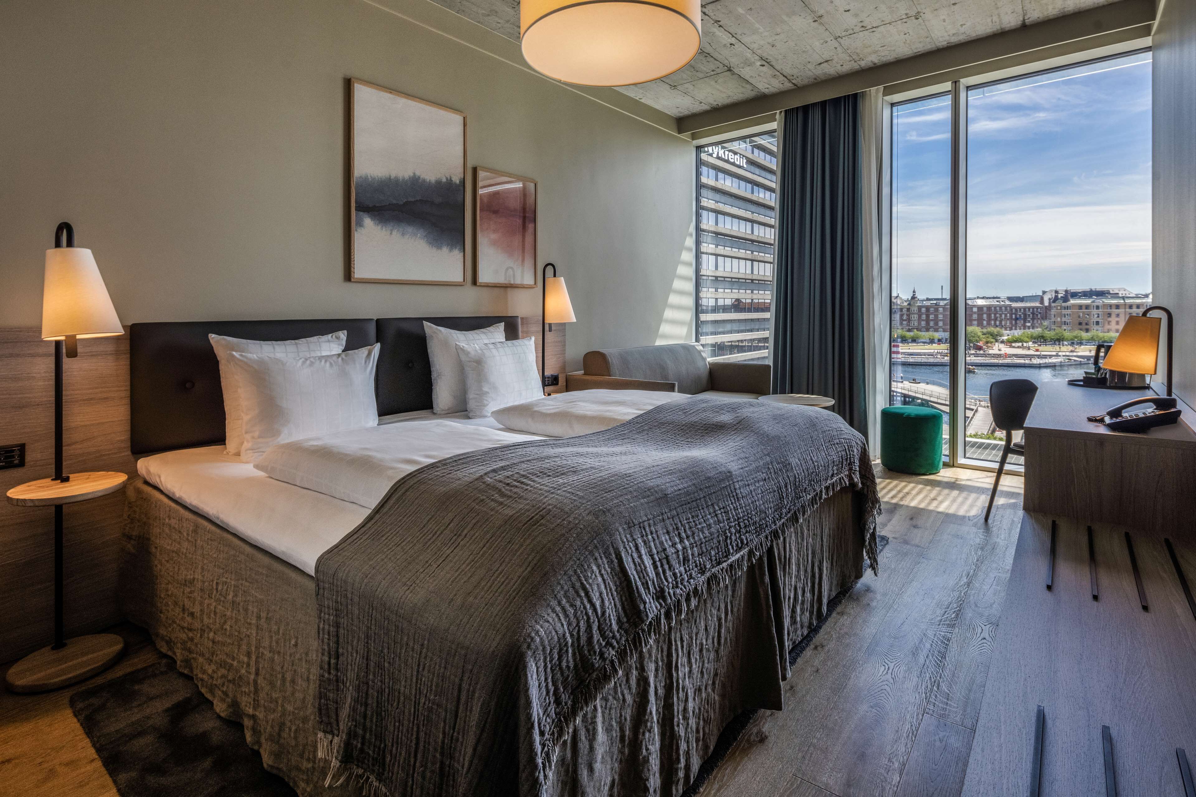 A modern hotel room with a large bed, two bedside tables with lamps, a desk, and a view of the city through large windows.