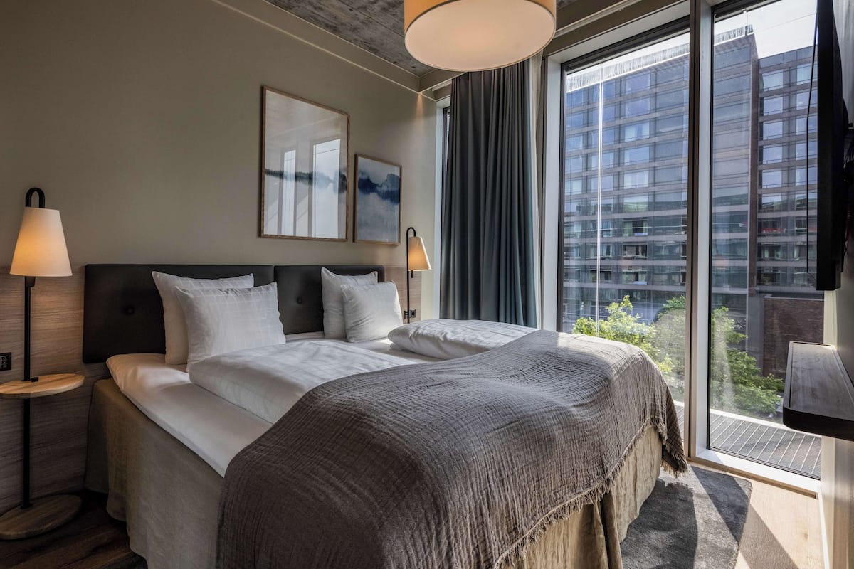 Une chambre d’hôtel moderne avec un grand lit, deux lampes de chevet, une vue sur la ville grâce à de grandes fenêtres du sol au plafond, et un panorama urbain visible à l’extérieur.