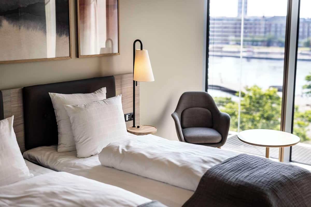 Une chambre d’hôtel avec un lit, une chaise, une petite table et une lampe. Une grande fenêtre offre une vue sur une rivière et des bâtiments.