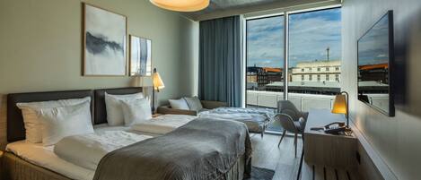 A hotel room with two beds, a desk, a chair, a large window with a city view, and artwork on the walls.