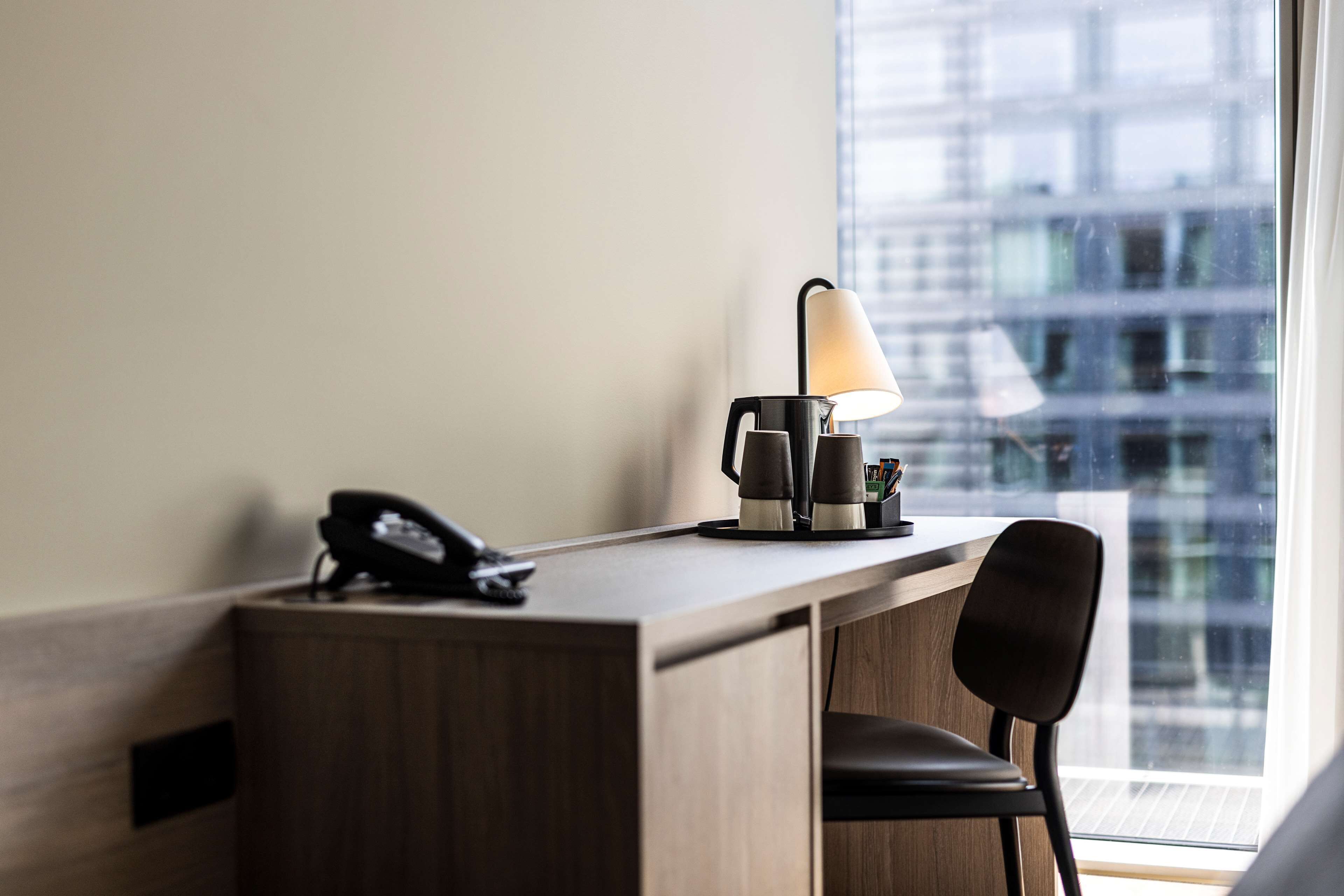 A hotel room with a wooden desk, a lamp, a telephone, and a view of a cityscape through a large window.