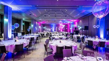 Un amplio salón de banquetes con mesas redondas dispuestas para cenar, iluminado con luces moradas y azules, y decorado con globos.