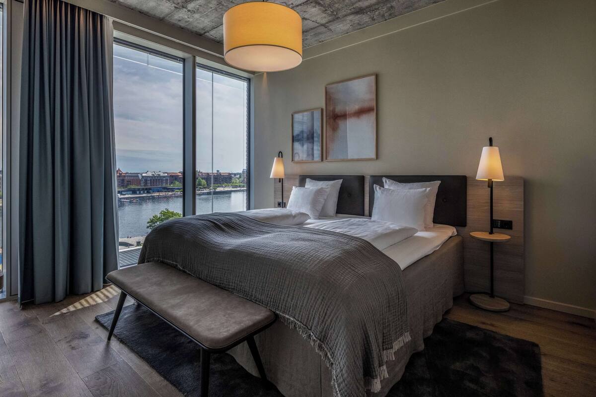 Une chambre d’hôtel moderne avec un grand lit, deux lampes de chevet, un banc et une vue sur le paysage urbain grâce à de grandes fenêtres.