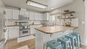 A kitchen with white cabinets, a wooden island with bar stools, a microwave, an oven, and a window with a view. - Family Home | 2 King Suites + Fire Pit | Walk to Breweries (Bend)
