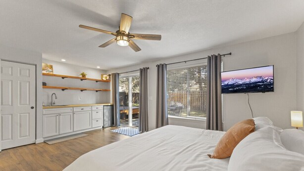 A bedroom with a bed, pillows, a TV, a ceiling fan, a kitchen area, and a sliding door to an outdoor space. - Family Home | 2 King Suites + Fire Pit | Walk to Breweries (Bend)