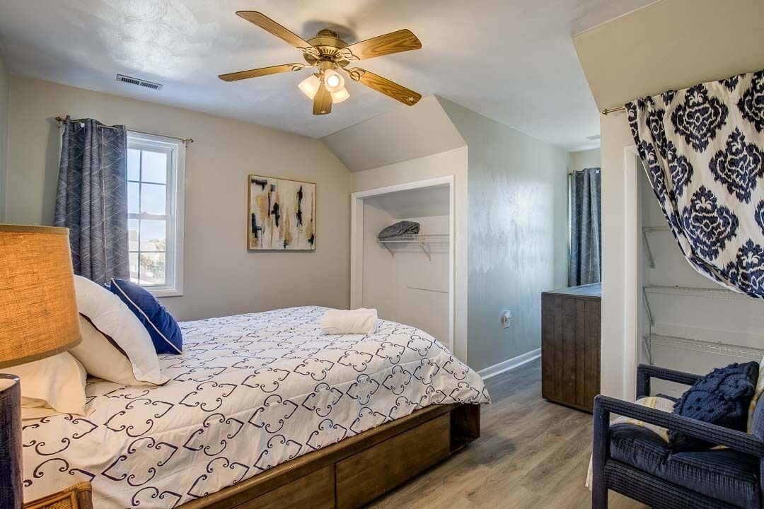 A bedroom with a bed, a ceiling fan, a window with curtains, a chair, and a door leading to another room.