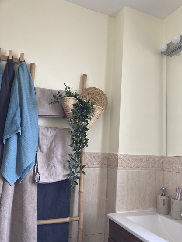 A bathroom with a wooden towel rack holding a blue towel and a beige towel, a woven basket with a plant, and a sink with two toothbrush holders.