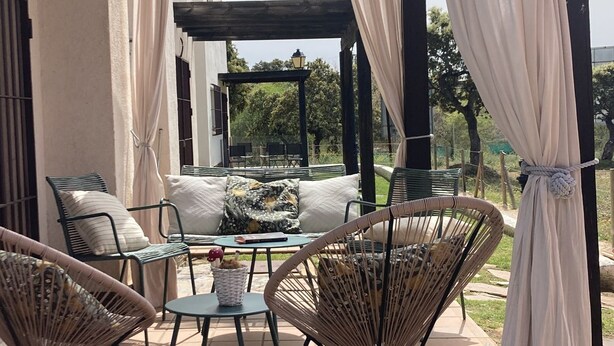 Une terrasse extérieure avec des meubles en osier, une table en verre et une pergola agrémentée de rideaux blancs.