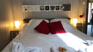 En pænt redt seng med hvide sengetæpper og røde puder, en sengetøjslampe og en hjerteformet dekoration på væggen.