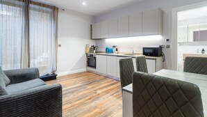 A modern kitchen with white cabinets, a microwave, and a sink. There is a dining area with a table and chairs. - Cosy one bedroom apartment in Highgate, close to Hampstead heath (London)