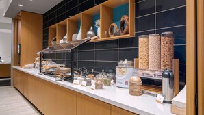 A buffet counter with various food items and containers, including jars of oatmeal and other cereals, a coffee machine, and a variety of dishes and utensils.