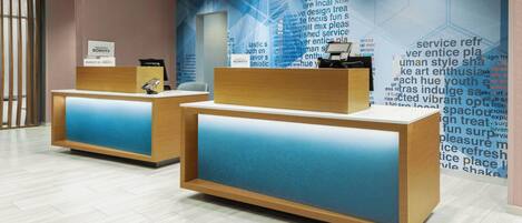 A hotel reception area with two wooden reception desks, a large wall with text and geometric patterns, and a modern interior design.