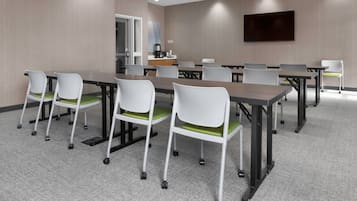 A conference room with a long table, white chairs with green cushions, a coffee station, and a flat-screen TV on the wall.