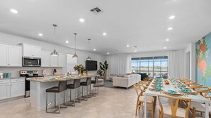 A modern kitchen with a central island, bar stools, and a dining area with a long table set for a meal. - 8BR Retreat | Sleep 20 | Theater | Huge Pool & Spa (Davenport)