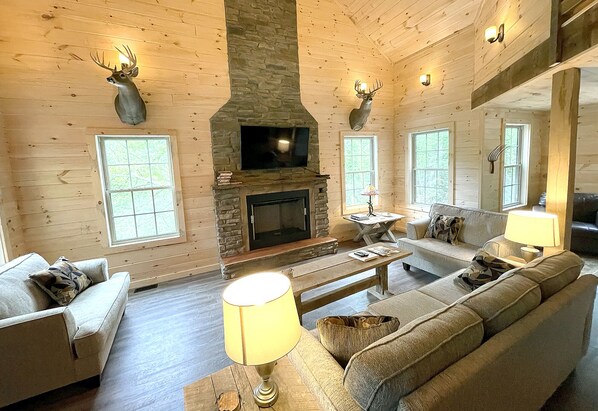 A log cabin interior with a stone fireplace, wooden walls, and mounted deer heads.