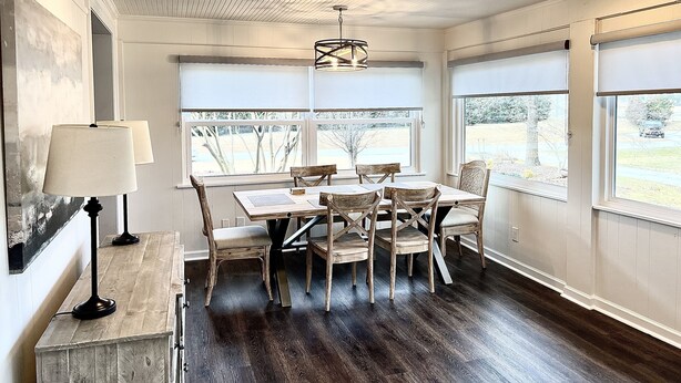 A dining area with a wooden table and chairs, a lamp, and large windows with views of trees and a road. - Centrally Located Cozy House,2 king beds,Game Room (Greensboro)