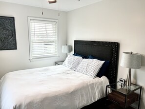 A neatly made bed with a white comforter and two pillows, a bedside table with a lamp, and a window with blinds.