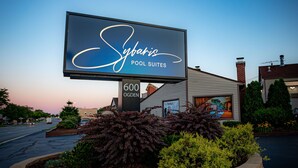 Schild für 'Sybaris Pool Suites' in der 600 Ogden, zeigt ein modernes Gebäude mit Backsteinfassade und eine begrünete Anlage mit Büschen.