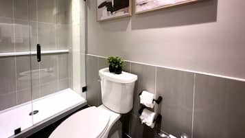 Un baño moderno con cabina de ducha de vidrio, inodoro con planta encima y dispensador de papel higiénico en la pared.