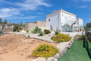 A white building with a stone wall, a staircase, and a garden with cacti and yellow flowers.